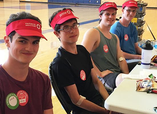 Four young men with light skin, wearing red American Red Cross visors, sitting at a table in a gymnasium. 他们穿着便服，贴着参加献血活动的贴纸.